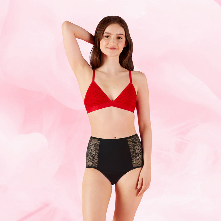 Against a light pink background, a woman smiles in a red bralette and the AltText.ai - High Waist Floral Moderate by intimawear by Bodyform™. The black high-waisted panties feature lace panels on the sides and TriTech Performance technology for style and comfort. She raises one hand to her head.